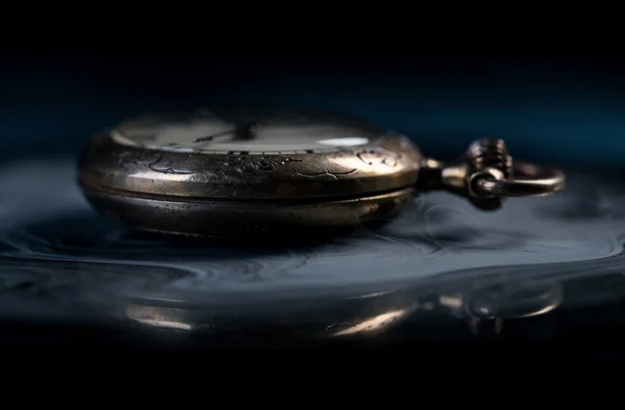 An antique pocket watch on dark glass, its reflection dissolving into soft mist
