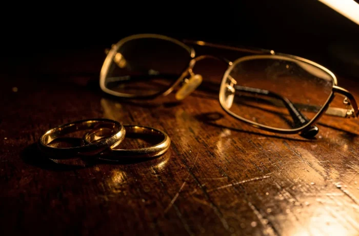 Two wedding rings on a dark wooden table beside folded reading glasses under warm lamplight