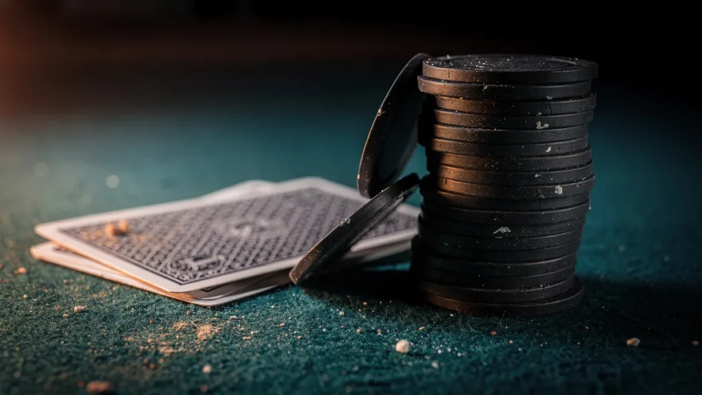 A stack of dark poker chips with two chips sliding off, beside face-down playing cards on worn teal felt under warm low light