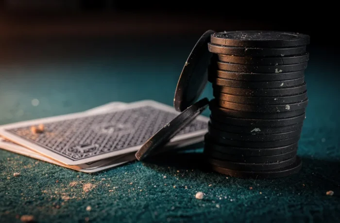 A stack of dark poker chips with two chips sliding off, beside face-down playing cards on worn teal felt under warm low light